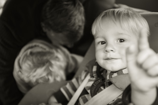Child sitting in a car seat, reaching toward the camera, while a father helps another child behind him