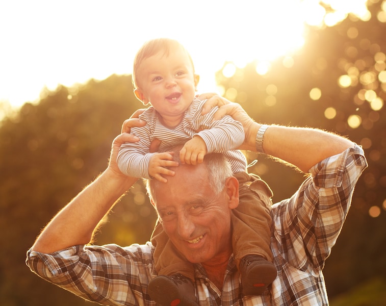 Older man holding a baby on his shoulders in front of a setting sun